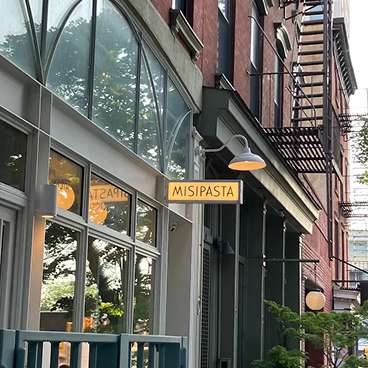 A street-side view of a restaurant exterior with a yellow sign reading MISIPASTA hanging from a brick building with large glass windows and outdoor wall lamps.