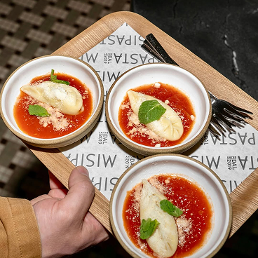 Three small bowls of pasta with red sauce, topped with a piece of white fish or potato, parmesan cheese, and a green mint leaf, arranged on a wooden tray held by a hand.